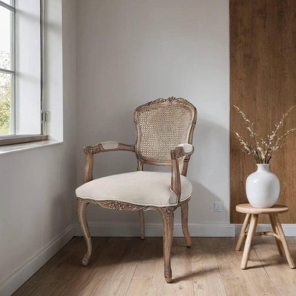 Vintage-style chair with white cushion next to a wooden stool with a vase and branches in a room with a window.