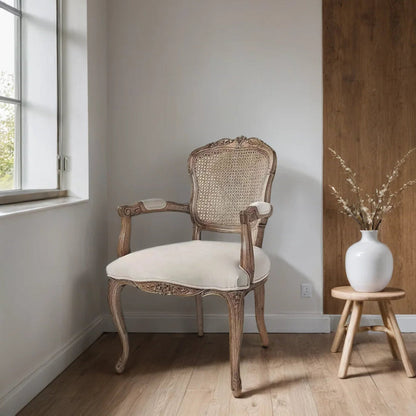 Vintage-style chair with white cushion next to a wooden stool with a vase and branches in a room with a window.