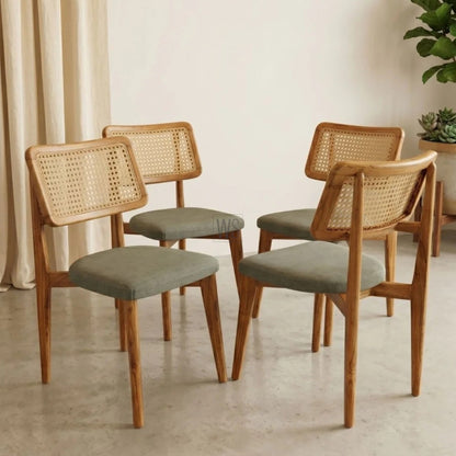 Four wooden chairs with wicker backrests and gray cushions in a room with light-colored walls and a plant.